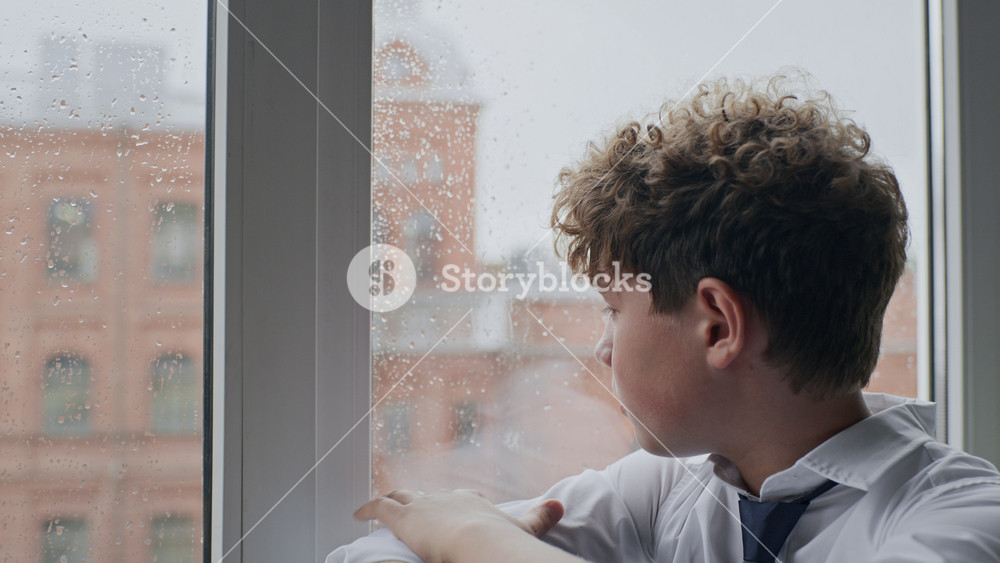 Caucasian teenager boy sitting on windowsill, lifting head to look through rainy window. Footage ideal for campaigns on youth mental health, school anxiety, and teenage depression