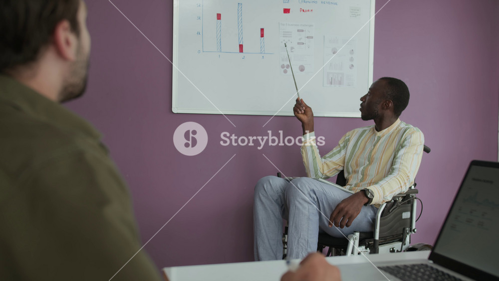 Tilt up shot of male worker listening to financial manager in wheelchair giving presentation at whiteboard at inclusive office