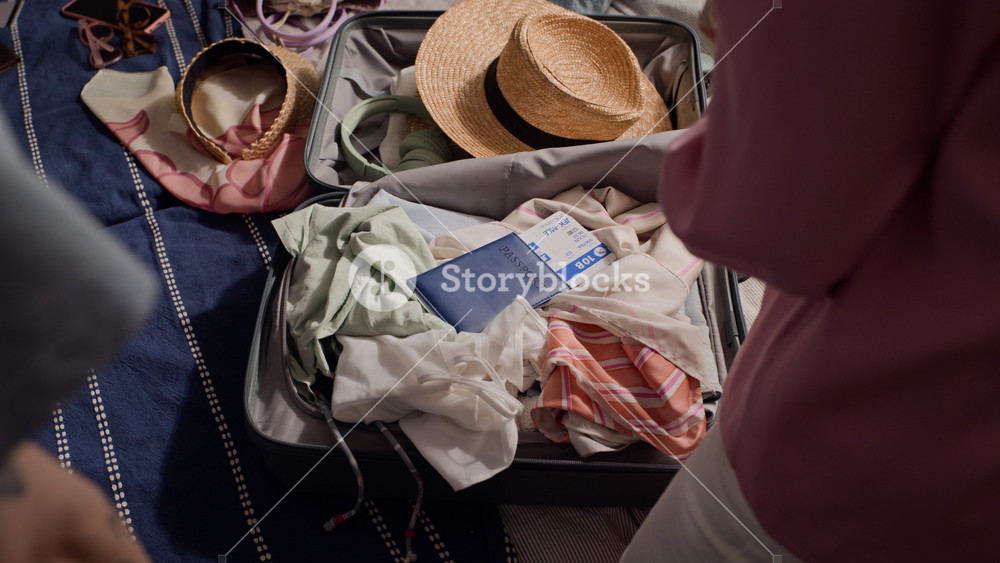 Back view of couple packing suitcase with summer clothes, straw hat, and passport on bed. Footage is perfect for travel agency ads and vacation planning campaigns