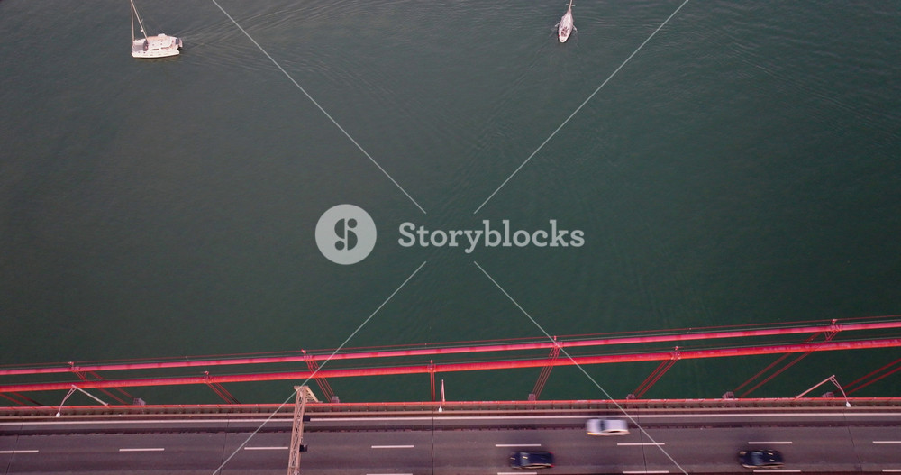 Top-down aerial drone shot of 25th April bridge with traffic and sailing boat and catamaran below. Lisbon and yacht lifestyle, yachting