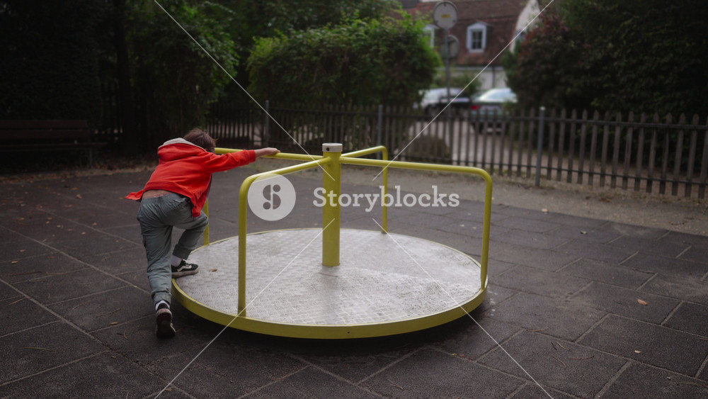 Child pushing yellow playground roundabout with focused energy in empty outdoor park setting as early autumn atmosphere and quiet residential backdrop frame moment of steady youthful motion