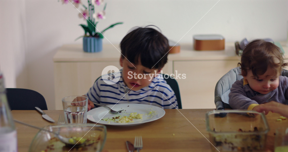 Mother shares family meal with two children at home dining table as restless older sibling shifts in his chair, capturing candid parenting and authentic everyday connection