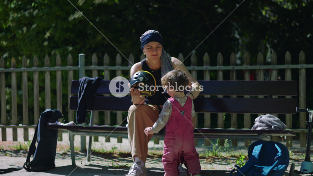 Mother undergoing chemotherapy placing helmet on toddler in park, headscarf parent ensuring child safety on bench, caring outdoor moment showing protection and resilience