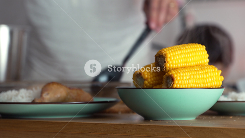 Close-up of freshly cooked corn on the cob served in a teal bowl, with blurred background of meal preparation symbolizing warmth, nourishment, and homemade food