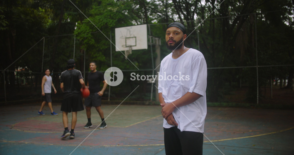 African descent man wearing durag with arms crossed on basketball court looking confident and focused while friends play behind, symbolizing leadership, resilience, and strength in teamwork