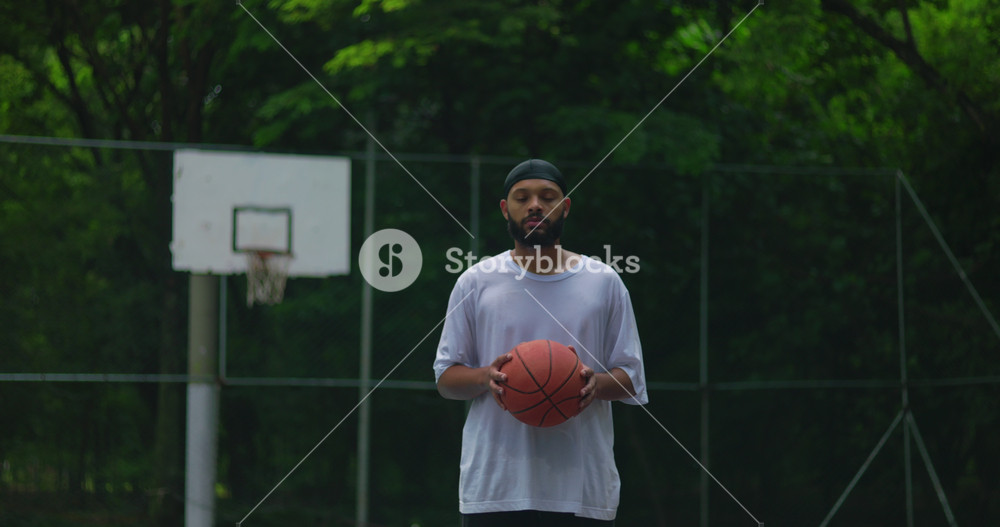 African American man standing on outdoor basketball court holding ball, looking straight ahead with calm focus, representing determination, confidence, and discipline in sports
