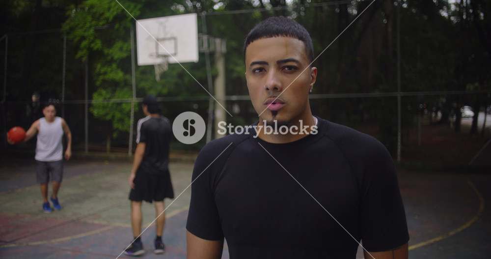 Focused young basketball player standing on outdoor court in black shirt, with teammates playing behind him, showing confidence, strength, and determination