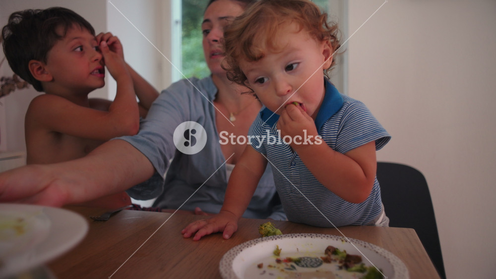 Toddler eating vegetables at table while mother and older brother sit nearby, concept of healthy habits, parenting guidance, family connection and childhood learning