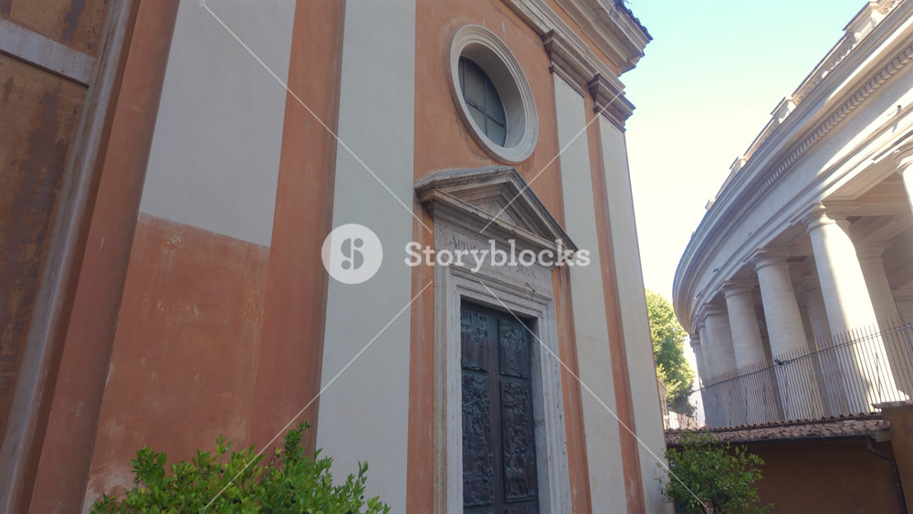 Facade Of Small Orange Church With Circular Window And Cross In Vatican City With Renaissance Details And Clear Blue Sky Above Historic Walls
