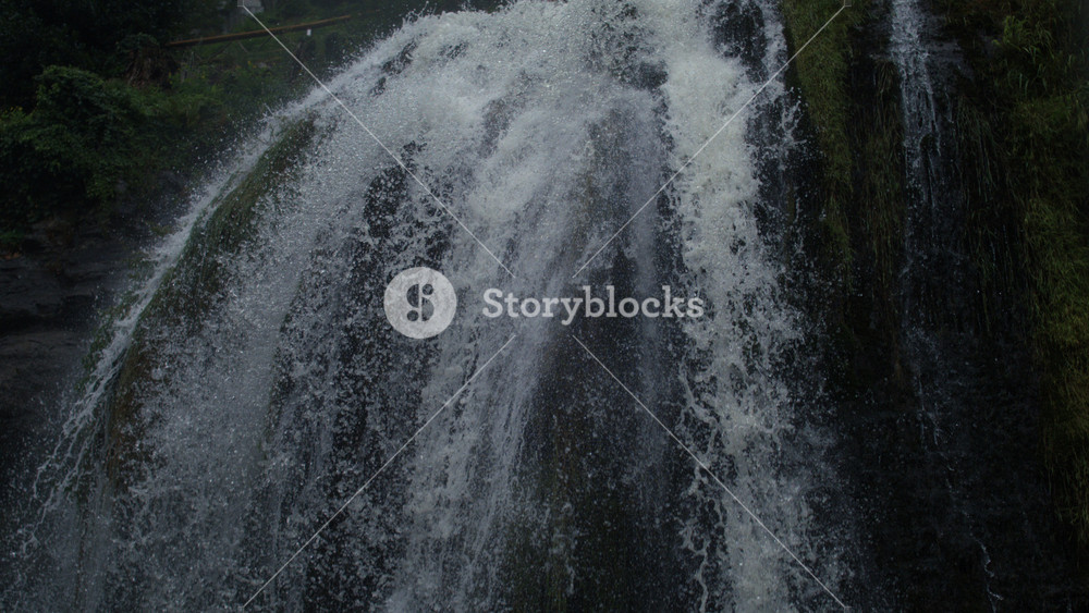Top of waterfall pouring down cliff surrounded by green vegetation, filmed in ultra slow motion with a high-speed camera, expressing grandeur and power of falling water