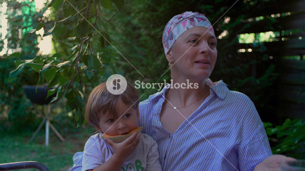 Mother with cancer holding toddler eating melon in a garden, nurturing outdoor family moment expressing resilience, connection and everyday joy through illness