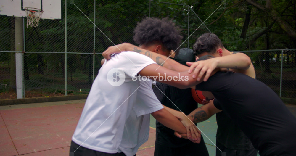 Group of Hispanic and African descent men huddling together on outdoor basketball court before game, arms over shoulders in team unity and pregame motivation