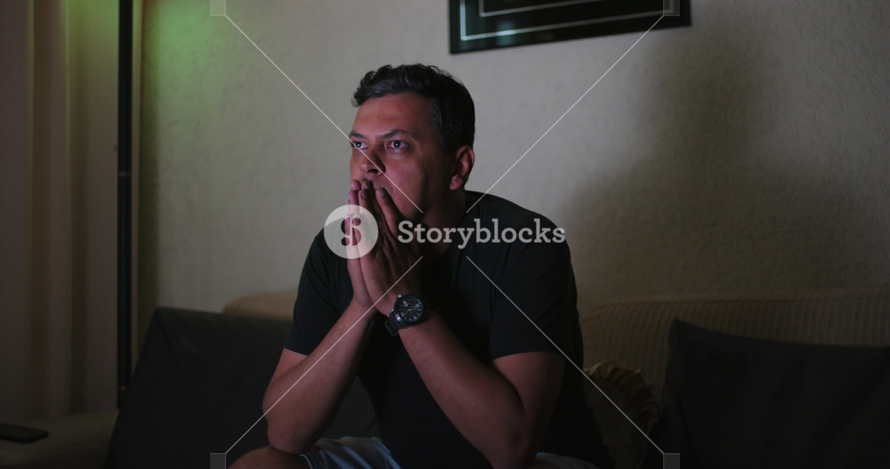 Man in deep suspense reacting to shocking TV content, clutching face with hands, wide-eyed disbelief, intense engagement, emotional tension, dramatic indoor moment