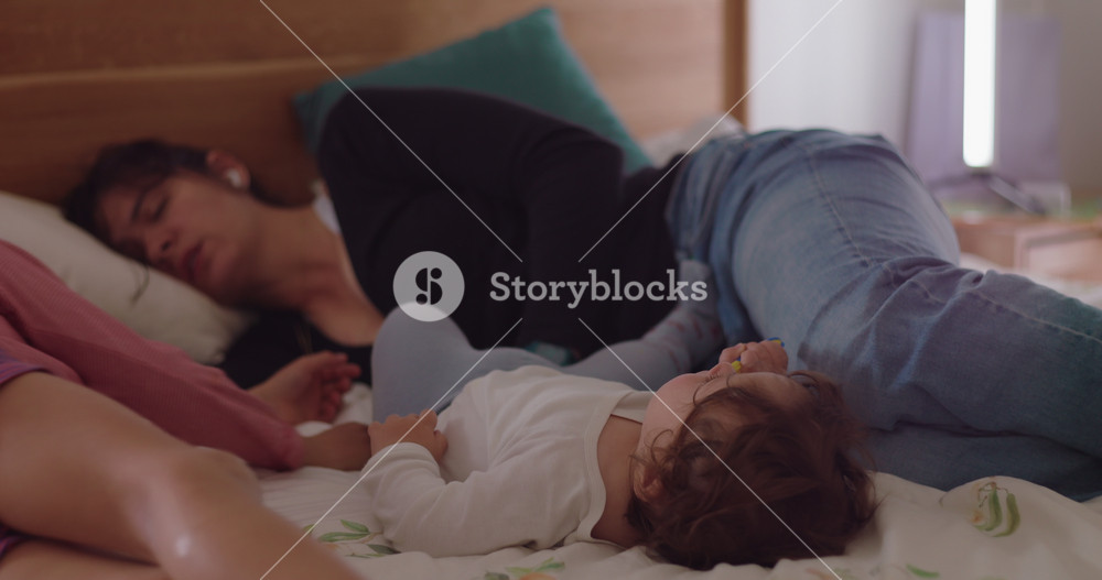 Active toddler rolls and climbs on bedroom bed beside older brother while exhausted mother sleeps and wakes overwhelmed, revealing maternal fatigue, family chaos and caregiving strain