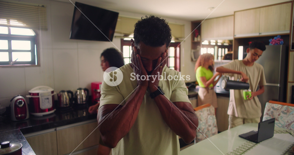 Father in foreground holding face in exhaustion and worry, overwhelmed by pressure, while calm and serene family in background carries on daily activities in the kitchen