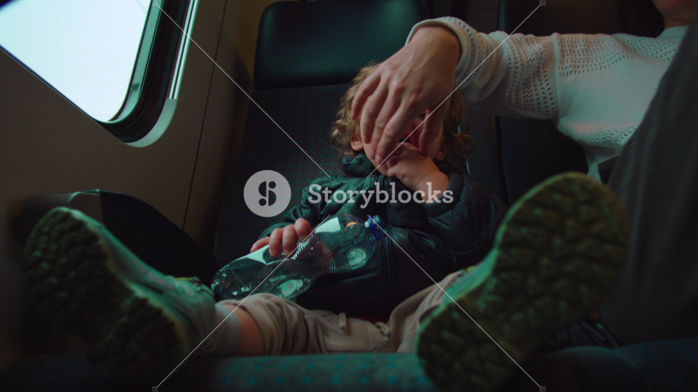 Toddler seated on train seat chewing plastic water bottle, playful expression and eye contact, candid travel moment showing curiosity, comfort, mischief, early childhood behavior