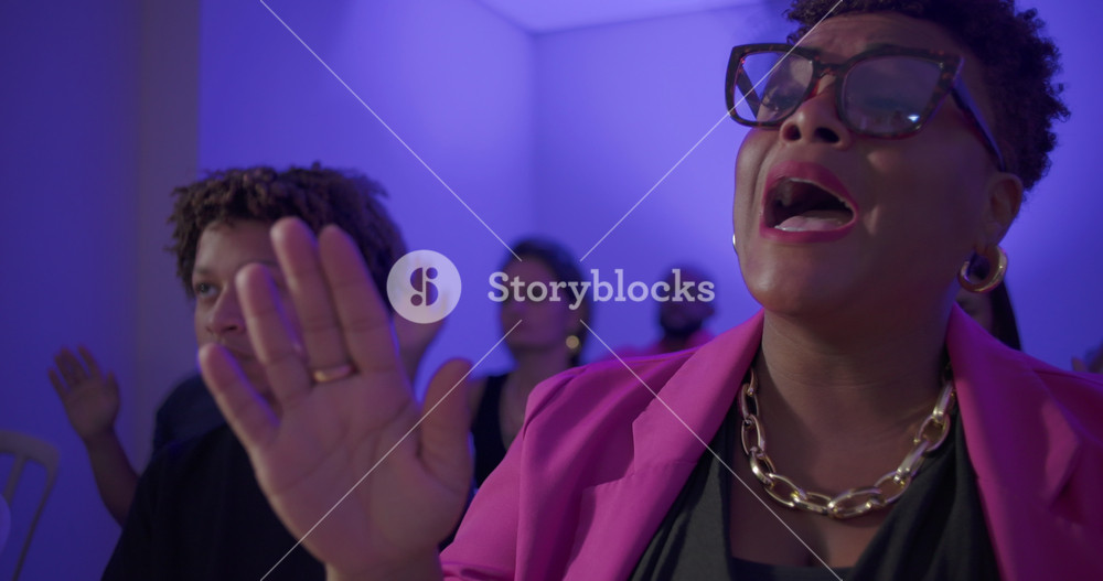 Close up of woman speaking and worshipping with hand raised during evangelical church service as others around her pray in the background