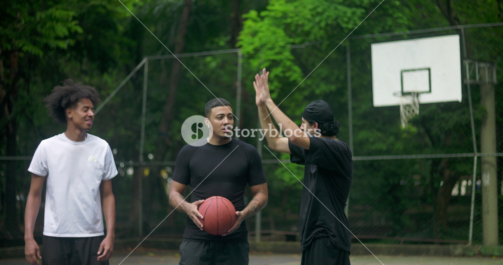 Three young men chatting and laughing on outdoor basketball court, enjoying friendship and relaxed conversation in casual urban sports setting surrounded by trees
