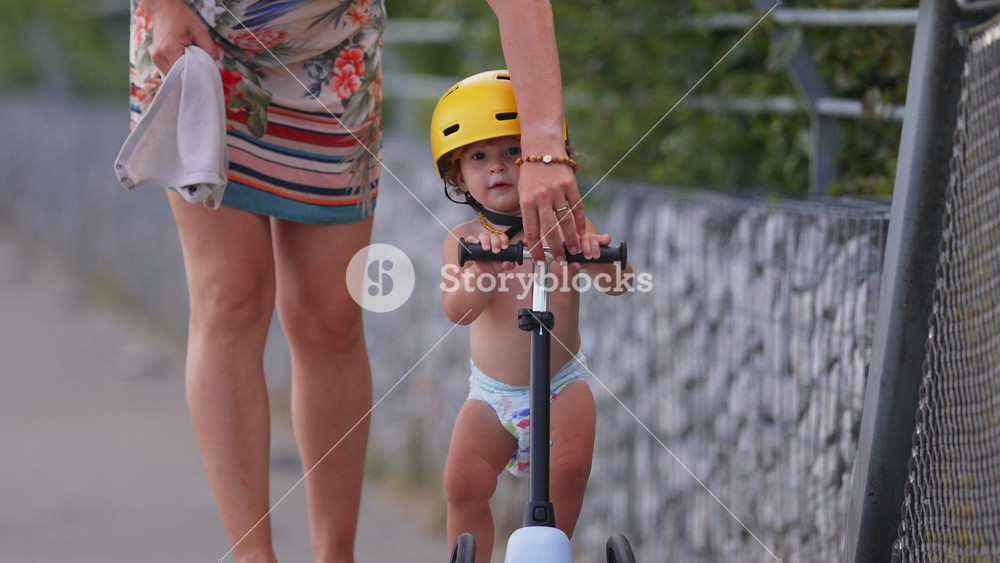 Close-up of toddler on scooter wearing yellow helmet while mother gently brakes handlebar, focusing on safety, protection, and early childhood learning outdoors