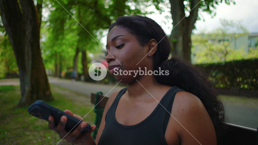 African American woman seated on park bench holding smartphone close to mouth and sending voice message creating natural and expressive communication moment in outdoor environment