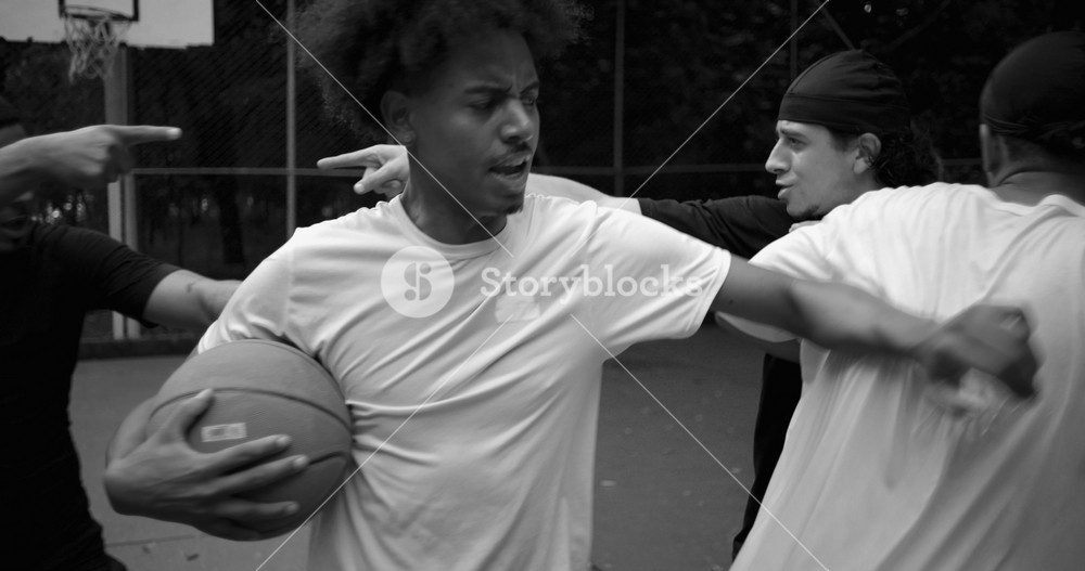 Hispanic and African descent men arguing on outdoor basketball court, yelling and pointing fingers in heated disagreement as teammate steps in to calm rising tension
