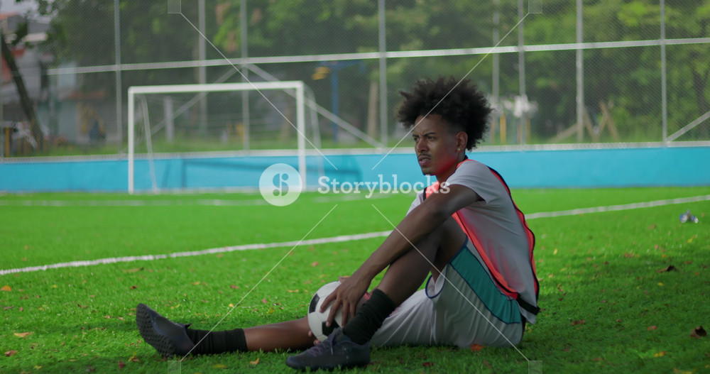 Brazilian man of African descent sitting on soccer field resting with ball between legs after match, looking away with thoughtful and slightly disappointed expression near goal net