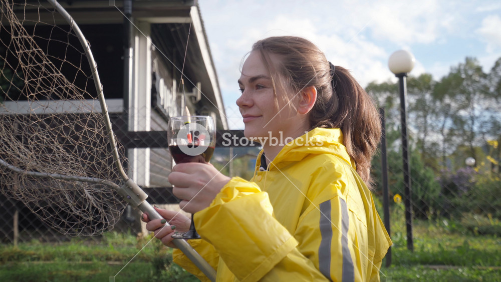 Caucasian woman in yellow raincoat watering plants holding wine glass while hose sprays water across suburban backyard near modern porch