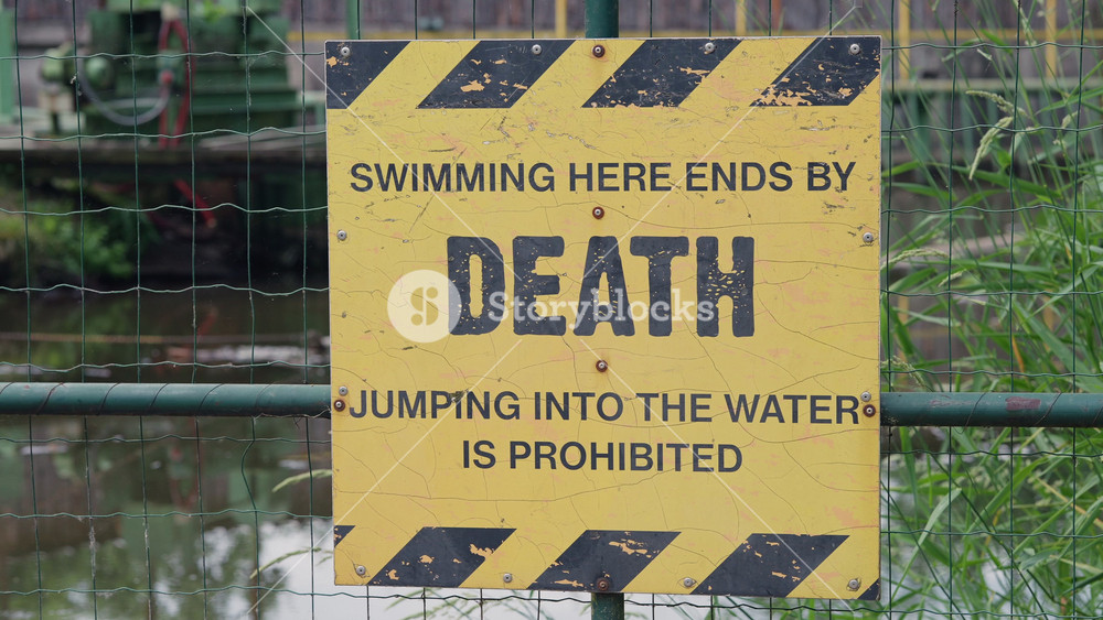 A close up of a weathered yellow warning sign on a metal fence, stating that swimming or jumping into the water is prohibited and may result