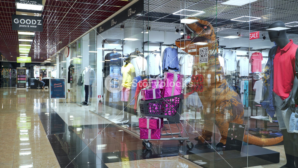Dinosaur costumed man leaving the shop with a cart full of presents. Orange T-Rex enters the children's clothes store in shopping mall.