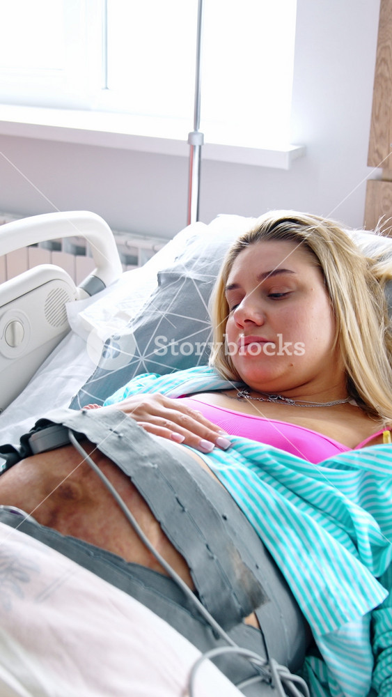 Blonde pregnant woman lies in bed with cardiotocography belt on the belly. Lady smiles to the camera. Top view. Vertical video