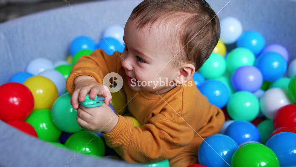 Joyful baby boy sitting in dry basin smiling adorably. Lovely child tries to climb out of the pool holding a pacifier in hand.