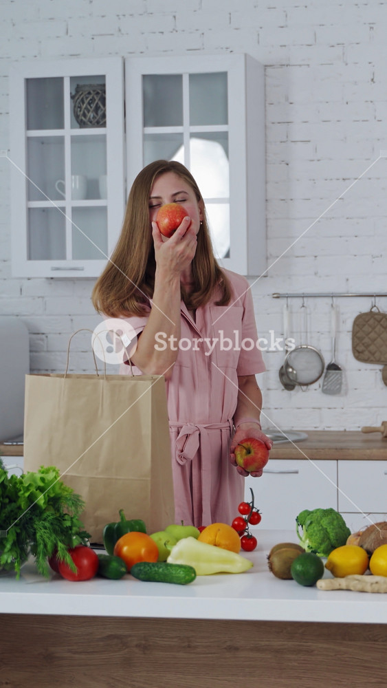 Happy young woman with fresh apples in the kitchen. Beautiful housewife holds and sniffs natural fruit on the background of healthy food on the table. Vertical video