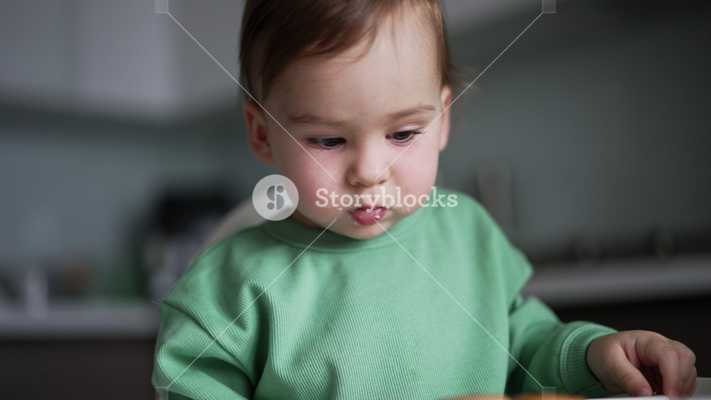 Pretty little baby with food around his mouth sits at feeding table. Mom's hand points at bagels in front of him and kid watches attentively.