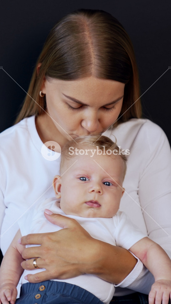 Long-haired blonde woman in white shirt holds her baby in hands. Loving mother kissing her child on the head. Black backdrop. Vertical video