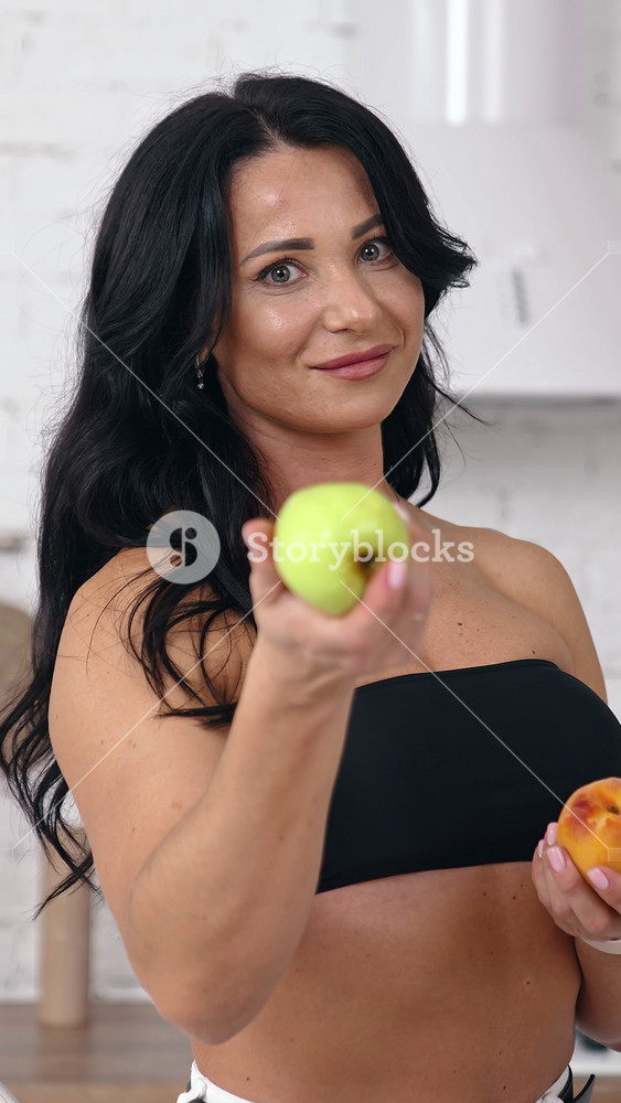Attractive female proposes fresh apples indoors. Portrait of a woman with bare shoulders and belly holding juicy fruit and showing on camera in the kitchen. Vertical video