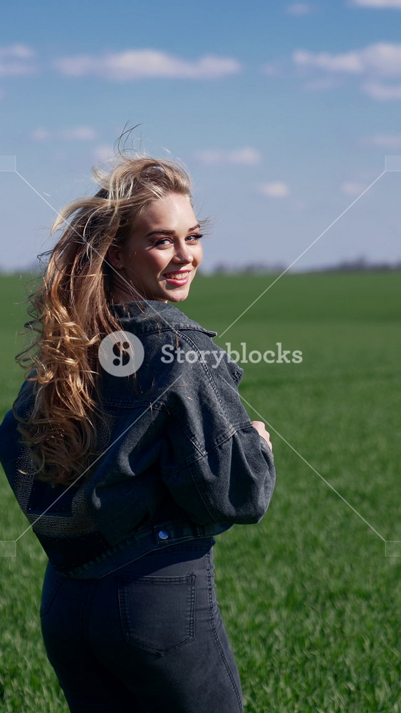 Girl is running on green field. Young woman with long fluttering hair in denim suit runs in nature and turning her cheerful face on camera. Rear view. Slow motion. Vertical video