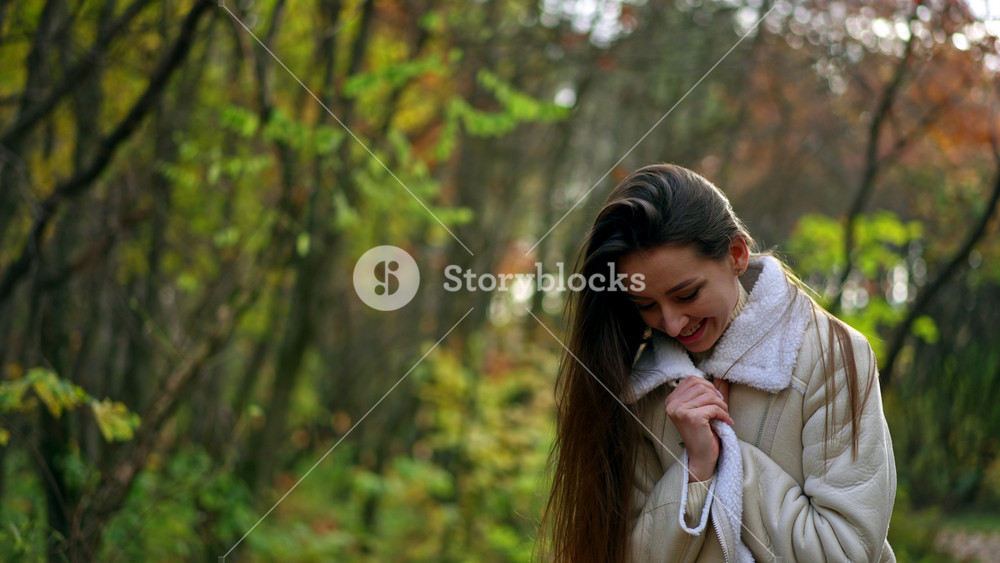 Dark-haired attractive lady in white jacket is in the autumn forest. Woman turns waving her long hair and smiling to the camera.