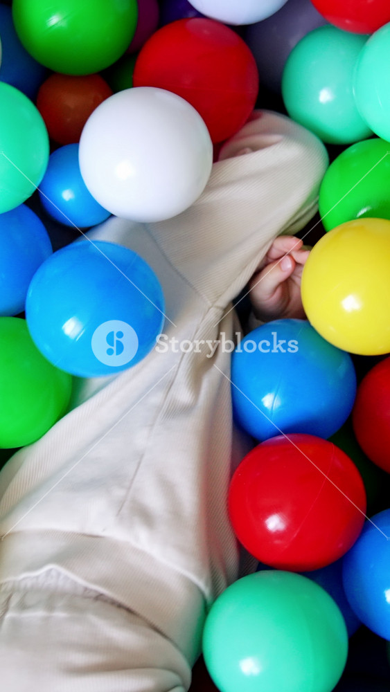 Baby boy lies hiding in the colorful balls in a dry pool. Toddler turns and sits in the basin taking one red ball in hands. Top view. Vertical video