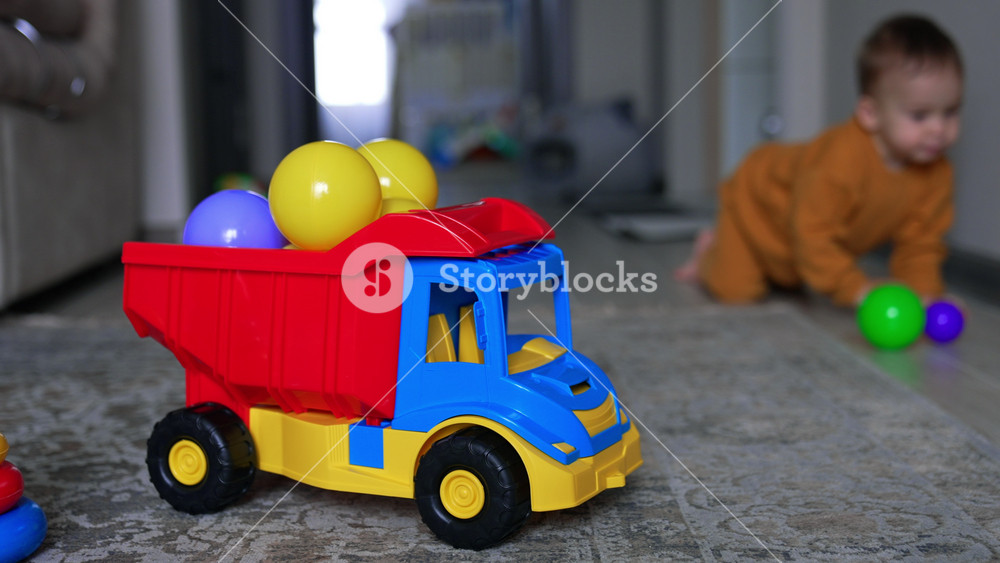 Colorful toy car with balls on the floor in the living room. Little baby appears in the room and crawls to the foreground. Blurred backdrop.