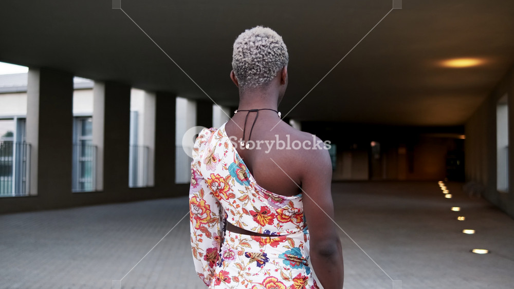 Side view of confident and smiling young African American androgynous person with short dyed hair and makeup in stylish dress standing on