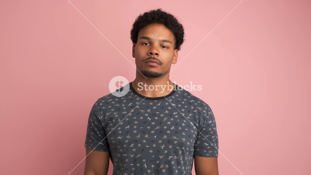 Portrait of upset young Brazilian male in t-shirt with afro hair showing thumb down while looking at camera standing over pink background