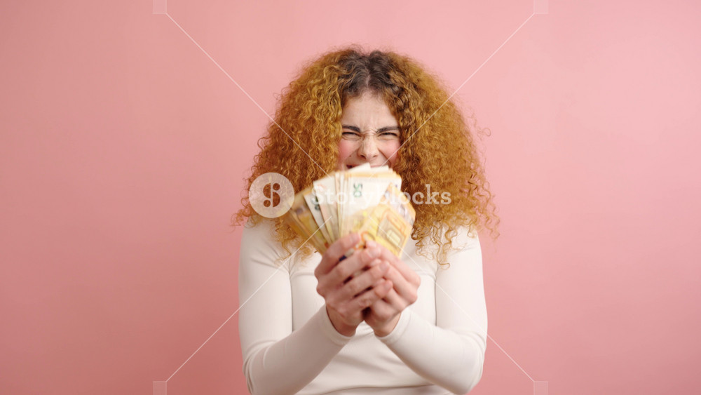 Portrait of joyful young woman with curly hair holding fan of euro banknotes expressing excitement and happiness against pink background
