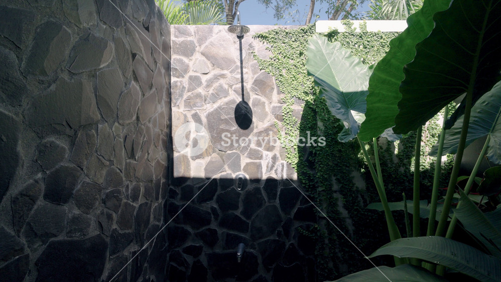 Sunlight revealing an outdoor shower with a stone wall in a luxury tropical resort. Lush green ivy and large jungle leaves surround the area