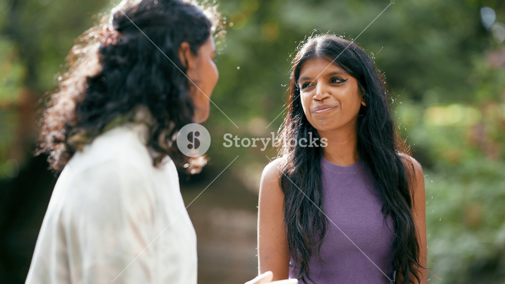 Two young south asian women standing in a park, talking and laughing together. Close up on the woman in the purple top listening to her