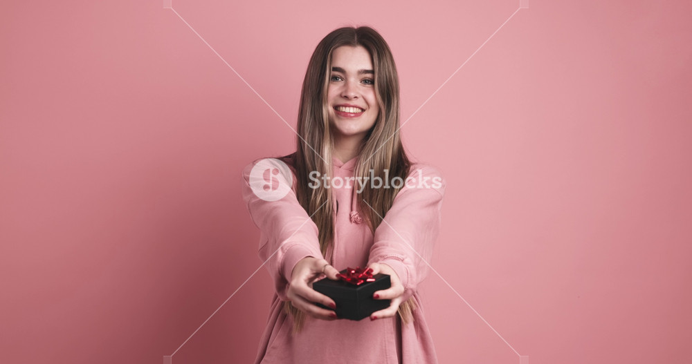 Portrait of smiling young female in trendy outfit with long dark hair giving surprise present box and looking at camera against pink