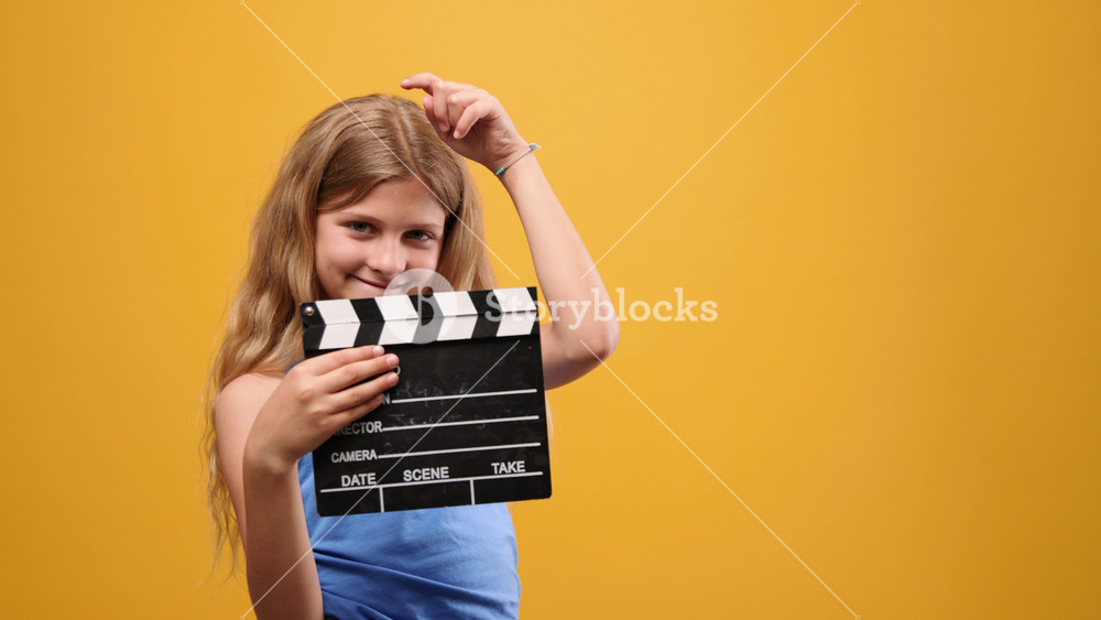Charming young girl with long blonde hair smiling and clapping a movie clapperboard, playing the role of a director on a yellow background