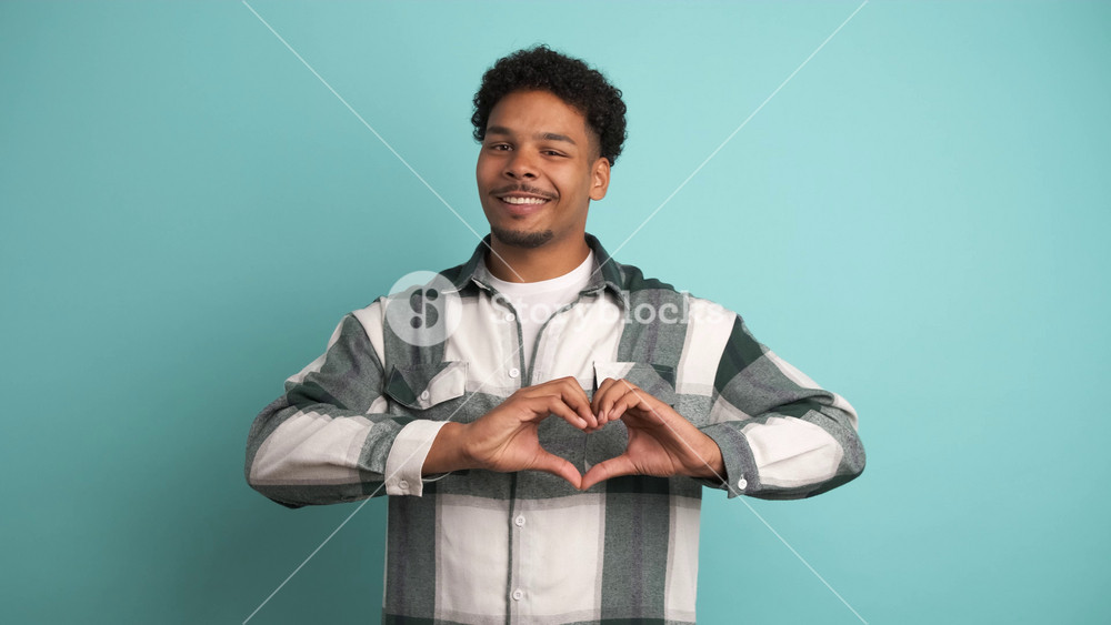 Portrait of smiling young Brazilian male in shirt with afro hair winking and showing heart gesture near chest while looking at camera over