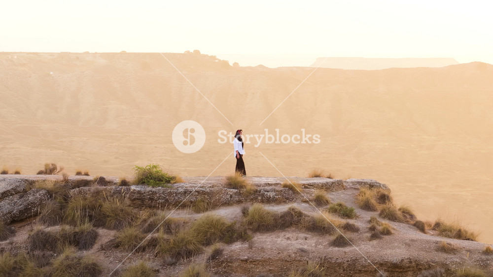 Aerial view of a woman in traditional clothing standing on the edge of a cliff. She is contemplating the vast arid landscape at sunset
