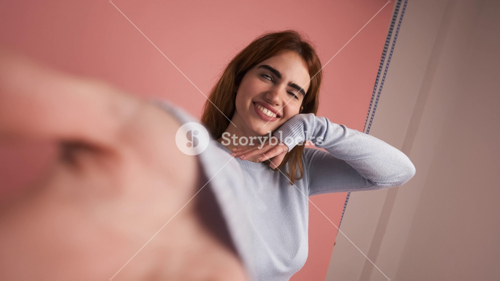 Portrait of smiling young female in casual clothes with dark hair making facial expressions while filming herself against peach color