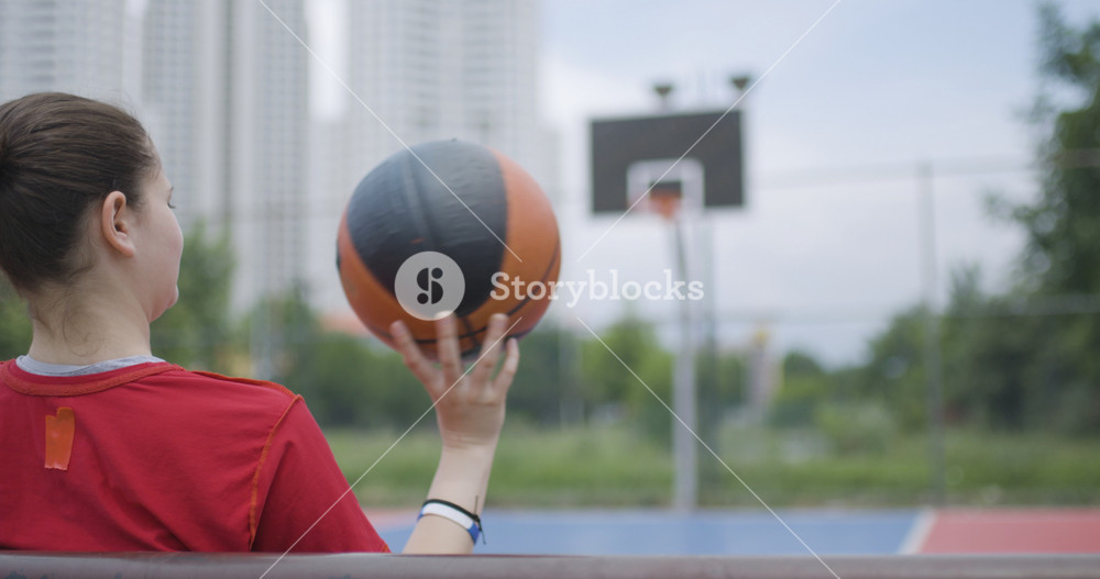 Female basketball player sitting on bench spinning ball in urban outdoor court with hoop in background